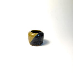 May include: A small, handcrafted ceramic vase with a unique color block design. The vase is a rounded cylinder shape, with a diagonal split of mustard yellow and dark blue-black. The interior is black.