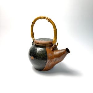 May include: A handcrafted ceramic teapot with a bamboo handle. The teapot is divided into black and brown sections, with a matching lid. The spout is brown, and the handle is made of curved bamboo. The teapot is designed for brewing tea.
