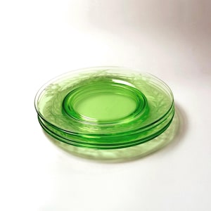 May include: A stack of three translucent light green glass plates featuring a delicate etched floral pattern along the rim. The circular plates have a smooth center and a raised edge, showcasing a vibrant lime green hue throughout the glass material.