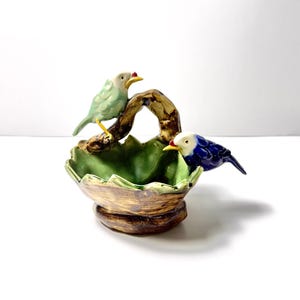 Peut inclure: Un bol en céramique avec un extérieur texturé marron et un intérieur vert, en forme de feuille. Une anse en forme de branche marron s'arque au-dessus du centre, avec un oiseau vert clair perché à gauche et un oiseau bleu foncé à droite.