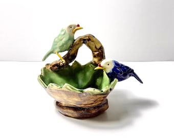 May include: A ceramic bowl featuring a brown textured exterior and a green interior, shaped like a leaf. A brown branch-like handle arches over the centre, with a light green bird perched on the left and a dark blue bird perched on the right side.