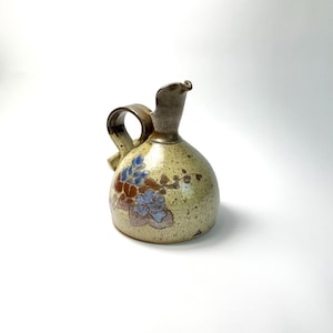 Könnte beinhalten: Kleine, handgefertigte Keramikkrug mit Henkel und Ausguss. Der Krug hat eine gesprenkelte, beige Grundfarbe mit einem floralen Design in Blau und Braun. Henkel und Ausguss sind dunkelbraun.