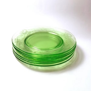 May include: A stack of four translucent light green glass plates featuring a delicate etched floral pattern along the outer rim. The plates have a smooth, circular design with a raised center section and a clear, glossy finish throughout.