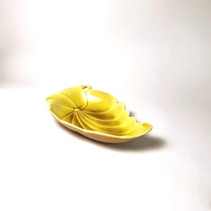Può includere: Un piatto in ceramica giallo brillante a forma di foglia o conchiglia stilizzata. L'oggetto presenta una finitura interna liscia e lucida con creste curve radianti che partono da un punto centrale, creando un motivo a ventaglio.
