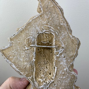 May include: A beige-colored, textured, irregular-shaped wall decor piece with a rectangular cutout in the center. The cutout has a rough, textured surface and a thin, silver-colored metal bar across the middle.