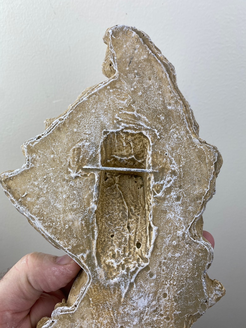 May include: A beige-colored, textured, irregular-shaped wall decor piece with a rectangular cutout in the center. The cutout has a rough, textured surface and a thin, silver-colored metal bar across the middle.
