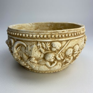 Pode incluir: Um vaso de cerâmica de cor creme com um design em relevo de querubins e motivos florais. O vaso tem uma borda de contas e um acabamento desgastado.