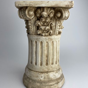 Può includere: Una colonna decorativa bianca con una base scanalata e una parte superiore dettagliata e ornata. La colonna è realizzata in un materiale testurizzato e ha un aspetto usurato e invecchiato.