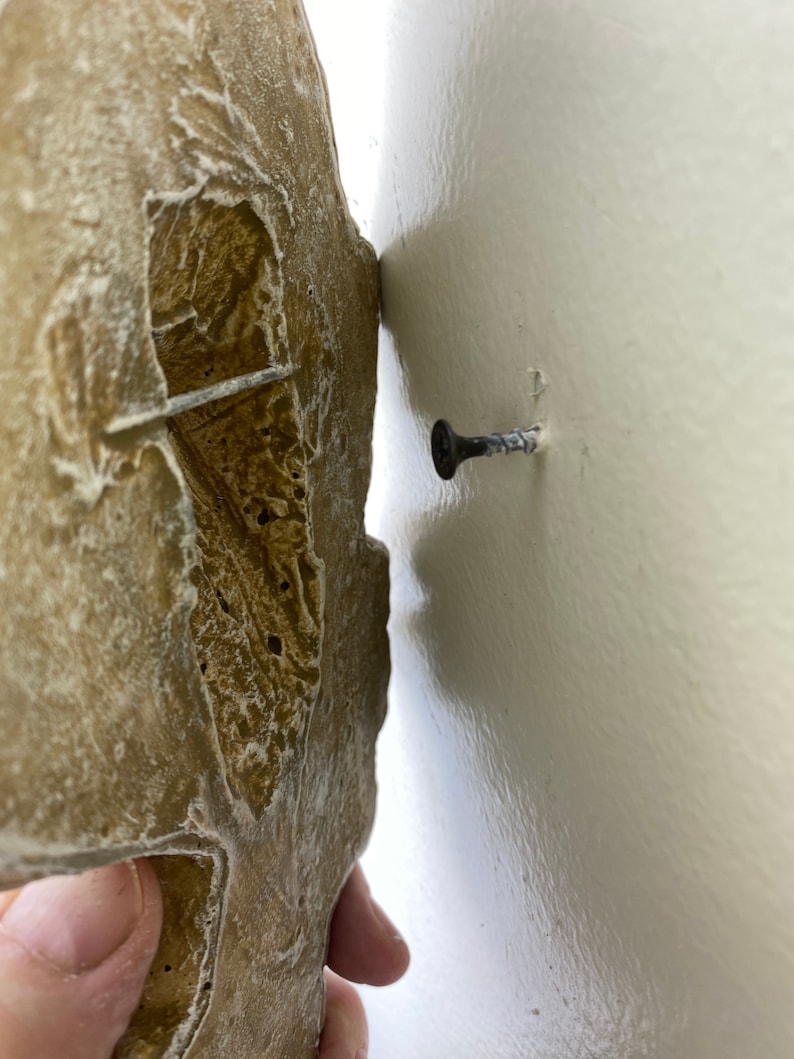 May include: A beige, textured wall decor piece with a hole in the center, being hung on a white wall with a screw.