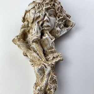 May include: A white and brown ceramic wall sculpture of a woman's face with leaves and branches surrounding her head.