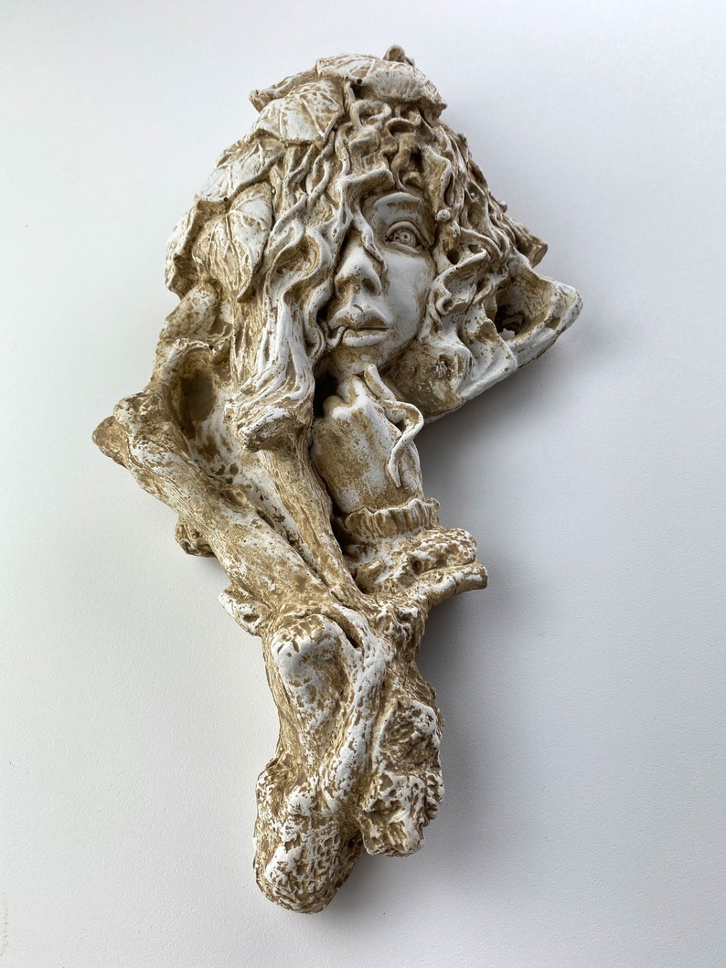 May include: A white and brown ceramic wall sculpture of a woman's face with leaves and branches surrounding her head.