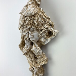May include: White ceramic wall sculpture of a woman's face with leafy hair and a tree trunk behind her. The sculpture has a rustic, earthy feel.