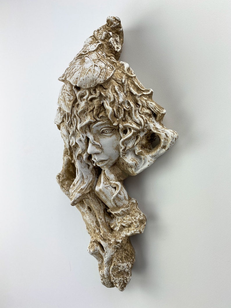 May include: White ceramic wall sculpture of a woman's face with leafy hair and a tree trunk behind her. The sculpture has a rustic, earthy feel.