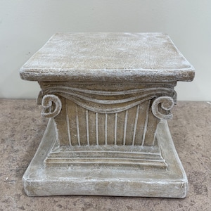 May include: A square pedestal stand with a weathered tan finish and white highlights. The base features a fluted column design with decorative scroll-like volutes at the top corners, supporting a flat, square top surface with a textured appearance.
