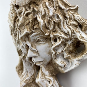 May include: A white and brown ceramic sculpture of a woman's face with detailed hair and a thoughtful expression.