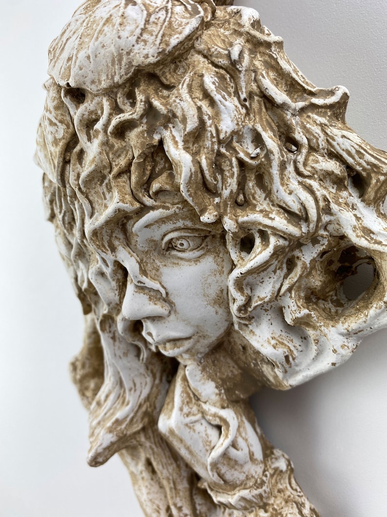May include: A white and brown ceramic sculpture of a woman's face with detailed hair and a thoughtful expression.