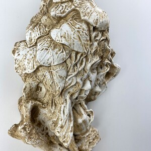 May include: A white ceramic sculpture of a woman's head with leaves and vines covering her hair. The sculpture has a distressed, aged look with brown and white paint.