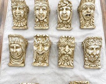 May include: A collection of 14 decorative wall brackets featuring various sculpted faces with intricate details. The brackets are made of a light-colored material, possibly plaster or ceramic, and have a distressed, antique finish.