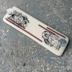 May include: A rectangular, cream-coloured decorative object featuring two black and white illustrations and two clear, faceted crystal knobs. Two parallel, dark red lines run down the centre of the object.