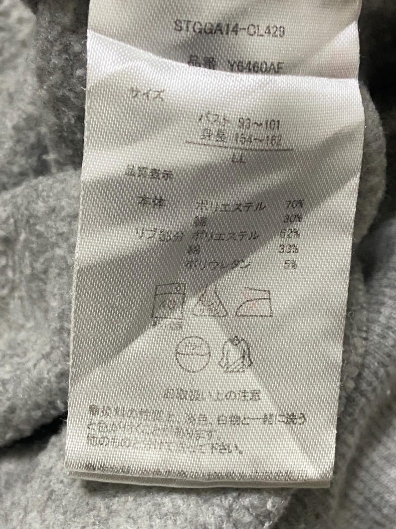 May include: A white tag with black text detailing the size, fabric composition, and care instructions for a grey sweatshirt. The tag includes the text "STGGA14-CL429", "Y6460AF", "バスト 93~101", "身長 154~162", "ポリエステル 70%", "30%", "リブ部分 ポリエステル 62%", "33%", "5%", and "他のものと分けて洗って下さい。"