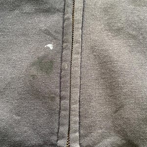 Pode incluir: Close-up de um casaco com capuz cinza escuro com z&iacute;per. O z&iacute;per corre verticalmente no centro, com o tecido de cada lado. Existem algumas manchas e uma pequena marca branca no tecido. A textura do material &eacute; vis&iacute;vel.