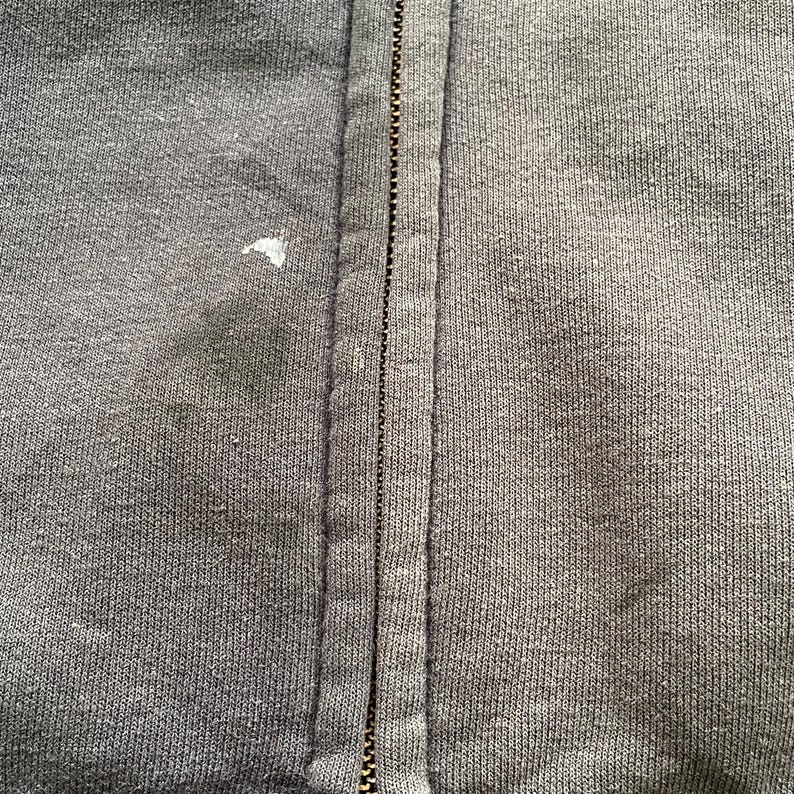 Pode incluir: Close-up de um casaco com capuz cinza escuro com z&iacute;per. O z&iacute;per corre verticalmente no centro, com o tecido de cada lado. Existem algumas manchas e uma pequena marca branca no tecido. A textura do material &eacute; vis&iacute;vel.