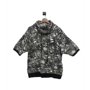 Op de afbeelding: Een zwart-witte hoodie met korte mouwen met een schedel- en kruisbeenderenprint. De hoodie heeft een hoge hals en een trekkoord.
