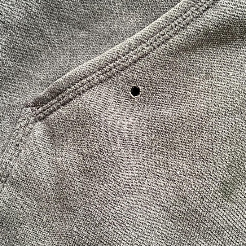 Pode incluir: Close-up de tecido cinza escuro com um buraco vis&iacute;vel. O tecido tem uma apar&ecirc;ncia texturizada e &eacute; costurado ao longo das bordas. A costura &eacute; em um tom de cinza mais claro, criando um contraste.