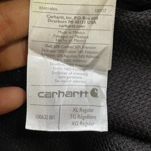 Pode incluir: Close-up de uma etiqueta Carhartt com texto. A etiqueta inclui o logotipo da Carhartt, endere&ccedil;o e informa&ccedil;&otilde;es de tamanho. A etiqueta tamb&eacute;m lista os materiais usados na pe&ccedil;a de vestu&aacute;rio, incluindo algod&atilde;o e poli&eacute;ster.