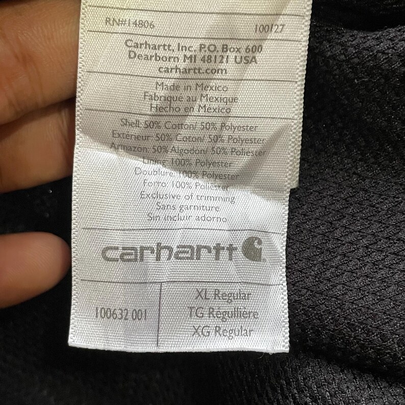 Pode incluir: Close-up de uma etiqueta Carhartt com texto. A etiqueta inclui o logotipo da Carhartt, endere&ccedil;o e informa&ccedil;&otilde;es de tamanho. A etiqueta tamb&eacute;m lista os materiais usados na pe&ccedil;a de vestu&aacute;rio, incluindo algod&atilde;o e poli&eacute;ster.