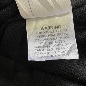 Pode incluir: Uma etiqueta branca com texto preto, avisando contra o uso de roupas isoladas perto de calor ou chamas. A etiqueta est&aacute; presa a um tecido preto, com o texto claramente leg&iacute;vel.