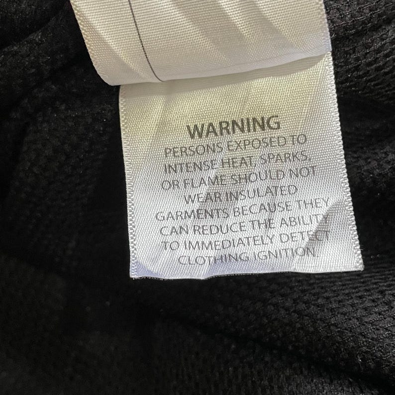 Pode incluir: Uma etiqueta branca com texto preto, avisando contra o uso de roupas isoladas perto de calor ou chamas. A etiqueta est&aacute; presa a um tecido preto, com o texto claramente leg&iacute;vel.