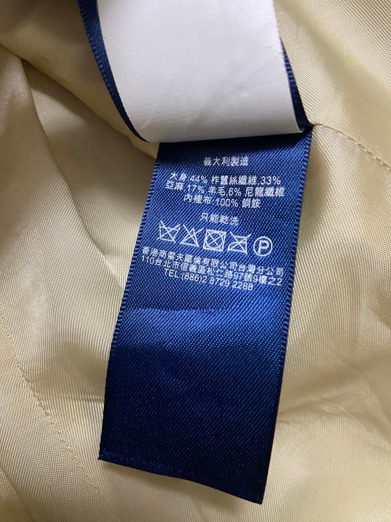 May include: Blue fabric care label with text in Japanese and English. The label states the fabric composition is 44% silk, 33% linen, 17% wool, 6% nylon, and 100% copper for the lining. The label also states "Made in Italy" and provides instructions for dry cleaning only.