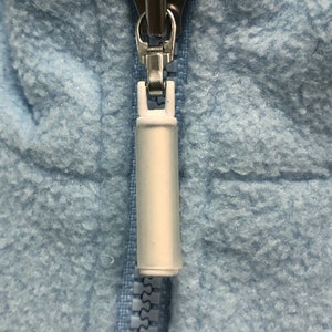 May include: A close-up of a light blue zipper with a white pull tab. The zipper is partially closed.