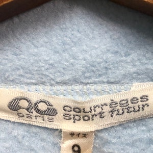 May include: A white tag with black text that reads "Courreges Sport Tour" and a logo of a stylized "C" with a circle around it. The tag also says "Paris" below the logo. A smaller white tag with black text says "サイズ" and "9" in a larger font.