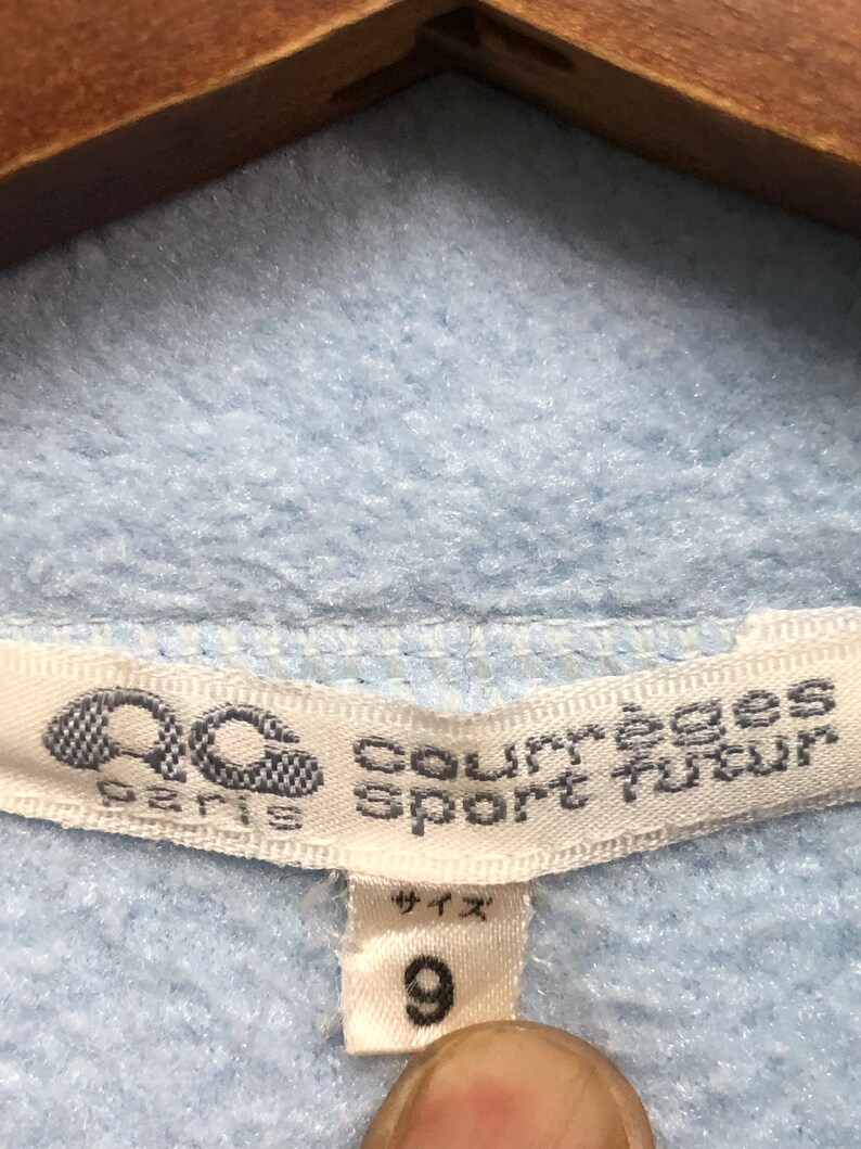May include: A white tag with black text that reads "Courreges Sport Tour" and a logo of a stylized "C" with a circle around it. The tag also says "Paris" below the logo. A smaller white tag with black text says "サイズ" and "9" in a larger font.