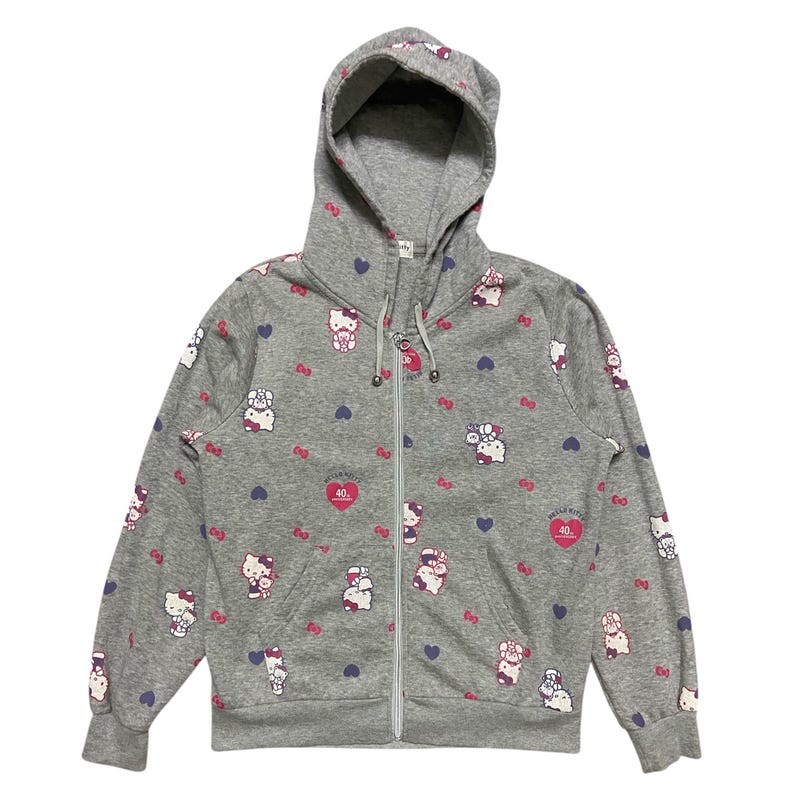 May include: A grey zip-up hoodie with a Hello Kitty print. The hoodie features a pink Hello Kitty with a red heart and the text "Hello Kitty 40th Anniversary" in a pink circle. The hoodie is covered in a pattern of Hello Kitty characters, hearts and bows.