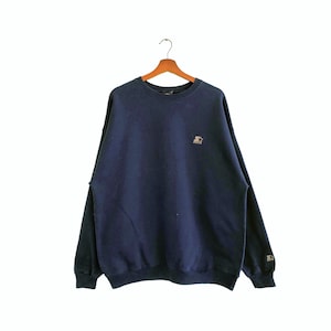 以下が含まれることがあります： 左胸に小さな刺繍ロゴが付いたネイビーブルーのスウェットシャツ。ロゴは黒い円の中に白い星があり、その下に「Starter」という文字が書かれています。