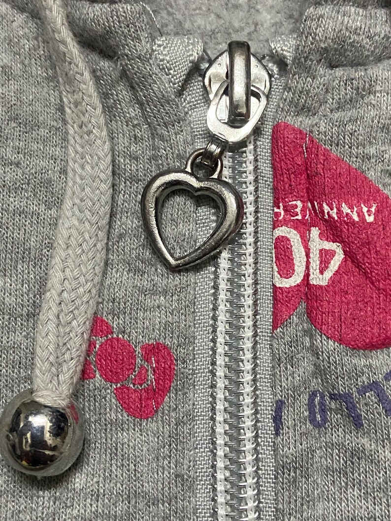 May include: A grey zip-up hoodie with a heart-shaped zipper pull. The hoodie has a pink heart with the text "40 ANNIVERSARY" printed on it.
