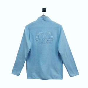 May include: A light blue fleece jacket with a zipper closure and a white embroidered logo that says "AC" and "Gourp&egrave;ges" with "Made in Italy" underneath.