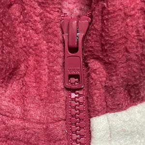K&ouml;nnte beinhalten: Nahaufnahme eines roten Rei&szlig;verschlusses an einer burgunderroten und wei&szlig;en Fleecejacke.