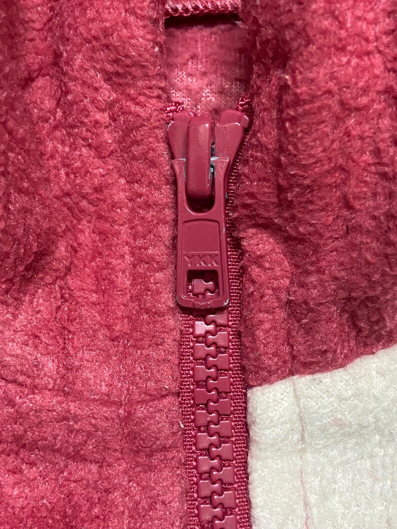 K&ouml;nnte beinhalten: Nahaufnahme eines roten Rei&szlig;verschlusses an einer burgunderroten und wei&szlig;en Fleecejacke.