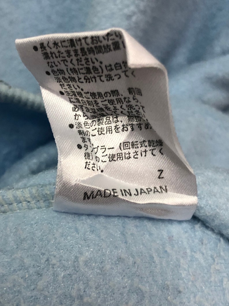 May include: White care tag with black text on a light blue background. The tag contains instructions for washing and drying the garment. The text reads: "Long soaking or leaving wet for long periods is not recommended.  Wash dark colors (especially dark colors) separately from light colors.  When using a washing machine, turn the garment inside out.  When using a dryer, use a low heat setting.  Do not use a dryer.  Made in Japan."