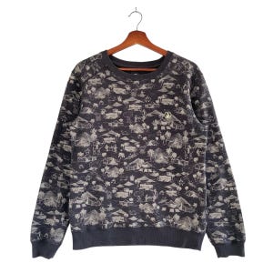 May include: Dark gray sweatshirt with a repeating white and gray tropical scene print. The design includes huts, palm trees, and figures. The sweatshirt has a crew neck and long sleeves. A small logo is visible on the upper chest.