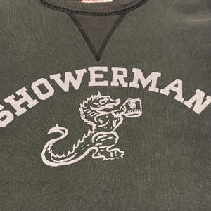 May include: Dark green sweatshirt with the word "SHOWERMAN" in large, light gray letters. Below the text is a light gray illustration of a dragon-like creature holding a mug. The sweatshirt has a black neckline.