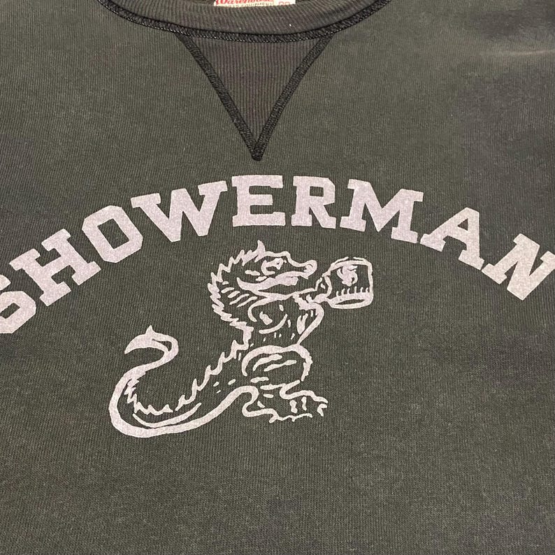May include: Dark green sweatshirt with the word "SHOWERMAN" in large, light gray letters. Below the text is a light gray illustration of a dragon-like creature holding a mug. The sweatshirt has a black neckline.