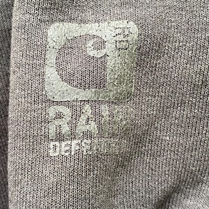 Pode incluir: Close-up de um tecido cinza com um logotipo desbotado e texturizado. O logotipo inclui uma forma de "C" estilizada e as palavras "RAW DEFENSE" em uma fonte desgastada. O tecido tem um padr&atilde;o sutil e tecido.