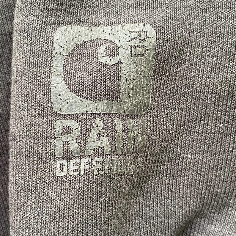 Pode incluir: Close-up de um tecido cinza com um logotipo desbotado e texturizado. O logotipo inclui uma forma de "C" estilizada e as palavras "RAW DEFENSE" em uma fonte desgastada. O tecido tem um padr&atilde;o sutil e tecido.