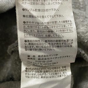 May include: A white care label with black text providing washing instructions for a garment. The label includes information about the material, care, and manufacturer. The text reads: "毛羽が付着することがあります。 洗濯ネットを使用して単独で洗濯をして下さい。 生地を損傷する場合がありますので、ファスナーは完全に閉じて洗って下さい。 タンブル乾燥はお避け下さい。 洗濯後は形を整えて干して下さい。 汗や水で湿った状態で放置したり摩擦すると、色移りすることがありますので注意して下さい。 また、着用時の摩擦により色移りすることがありますので、注意して下さい。 素材の特性上、着用時の摩擦や洗濯により、表面の毛羽立ち、毛玉が発生することがあります。 特に毛玉の目立つ場合には毛布取り器で取り除いて下さい。 著作 株式会社サンリオ 発売元 株式会社カイタックファミリー FOR SALE IN JAPAN ONLY 0120-66-2240 ●ひも止めなどが付いている商品は、自宅等のドア・自転車・遊具などに絡まる危険性があります。 ひもが長く下がる場合は、結んで短くするなど、絡まないようにご注意下さい."