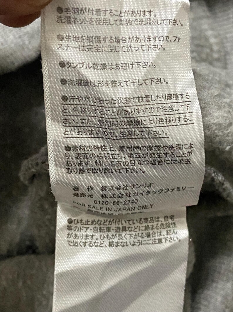 May include: A white care label with black text providing washing instructions for a garment. The label includes information about the material, care, and manufacturer. The text reads: "毛羽が付着することがあります。 洗濯ネットを使用して単独で洗濯をして下さい。 生地を損傷する場合がありますので、ファスナーは完全に閉じて洗って下さい。 タンブル乾燥はお避け下さい。 洗濯後は形を整えて干して下さい。 汗や水で湿った状態で放置したり摩擦すると、色移りすることがありますので注意して下さい。 また、着用時の摩擦により色移りすることがありますので、注意して下さい。 素材の特性上、着用時の摩擦や洗濯により、表面の毛羽立ち、毛玉が発生することがあります。 特に毛玉の目立つ場合には毛布取り器で取り除いて下さい。 著作 株式会社サンリオ 発売元 株式会社カイタックファミリー FOR SALE IN JAPAN ONLY 0120-66-2240 ●ひも止めなどが付いている商品は、自宅等のドア・自転車・遊具などに絡まる危険性があります。 ひもが長く下がる場合は、結んで短くするなど、絡まないようにご注意下さい."
